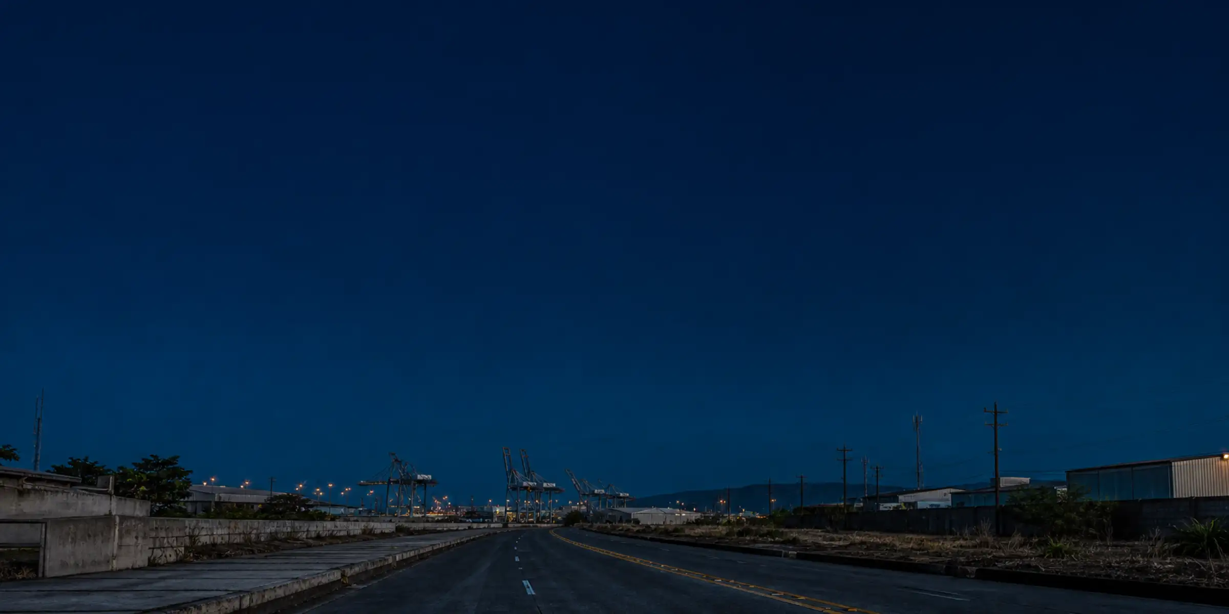 Una carretera vacía avanza al amanecer hacia las grúas de un puerto, entre bodegas y postes de luz.